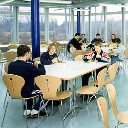 Verglaster Speisesaal; ein Tisch mit drei essenden Personen im Vordergrund und weiteren Tischen und Personen im Hintergrund